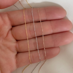 May include: Close-up of four delicate chain necklaces in gold, silver, and rose gold colors. The chains have a fine, rectangular link design, and are displayed against a soft, neutral background.