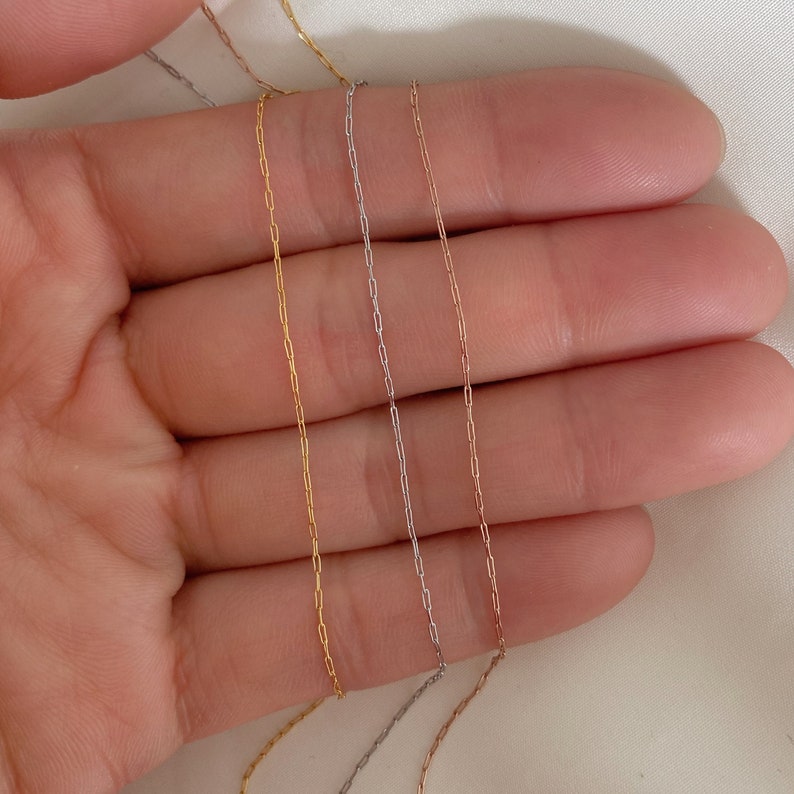 May include: Close-up of four delicate chain necklaces in gold, silver, and rose gold colors. The chains have a fine, rectangular link design, and are displayed against a soft, neutral background.