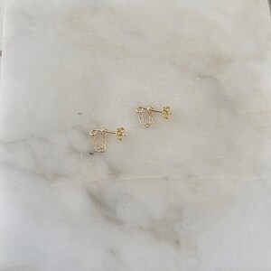 Puede incluir: Un par de pendientes de cadena dorados. Cada pendiente presenta una peque&ntilde;a cadena que cuelga de un poste. Los pendientes se muestran sobre una superficie de m&aacute;rmol blanco y gris.