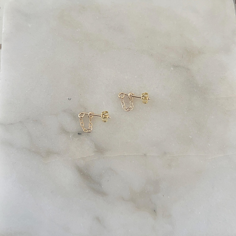 Puede incluir: Un par de pendientes de cadena dorados. Cada pendiente presenta una peque&ntilde;a cadena que cuelga de un poste. Los pendientes se muestran sobre una superficie de m&aacute;rmol blanco y gris.