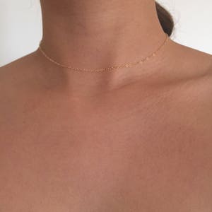 May include: A delicate gold chain necklace, worn as a choker, rests on a person's neck. The chain is thin and simple, with a subtle shine.