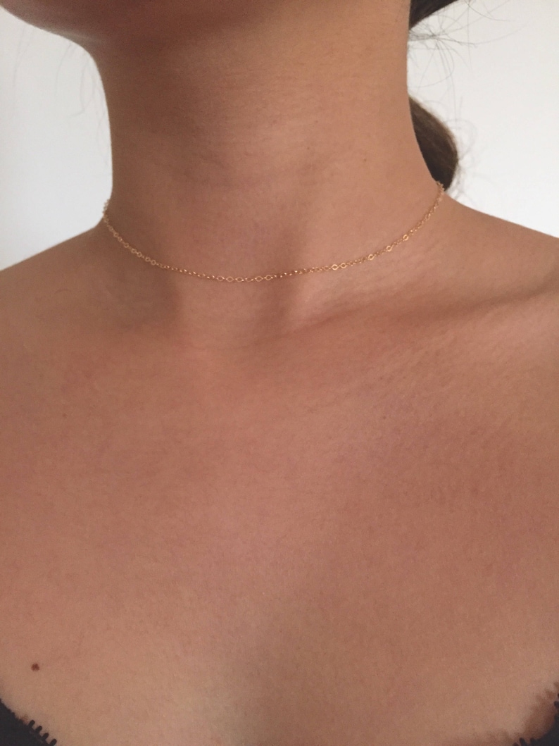 May include: A delicate gold chain necklace, worn as a choker, rests on a person's neck. The chain is thin and simple, with a subtle shine.