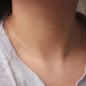 Könnte beinhalten: Eine zarte silberne Kettenkette um den Hals einer Person. Die Kette ist dünn und hat einen dezenten Glanz.