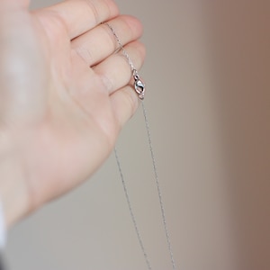 May include: A delicate silver necklace with a fine chain and a small clasp. The necklace is held in a hand, showcasing its simple elegance and design. The chain appears to be approximately 18 inches long.