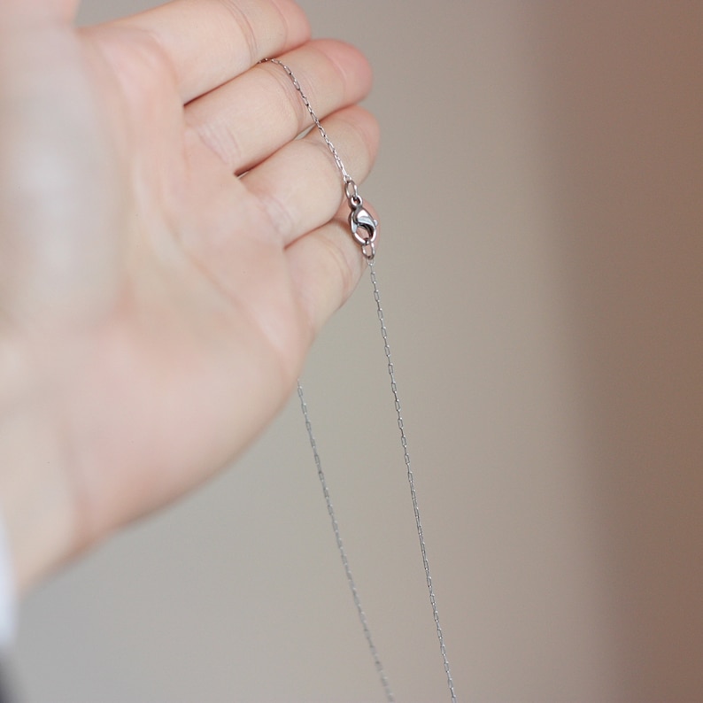 May include: A delicate silver necklace with a fine chain and a small clasp. The necklace is held in a hand, showcasing its simple elegance and design. The chain appears to be approximately 18 inches long.