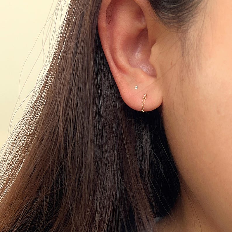 Puede incluir: Primer plano de una oreja adornada con dos pendientes dorados. Uno es un peque&ntilde;o perno y el otro es un pendiente de cadena que cuelga con gracia. Los pendientes a&ntilde;aden un toque de elegancia a la oreja.