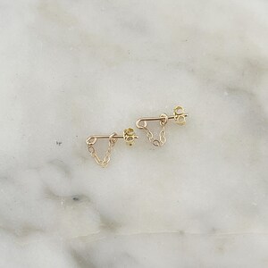 Puede incluir: Un par de pendientes de cadena dorados. Cada pendiente tiene un poste con una cadena que cuelga en forma de V. Los pendientes se muestran sobre una superficie de m&aacute;rmol blanco y gris.