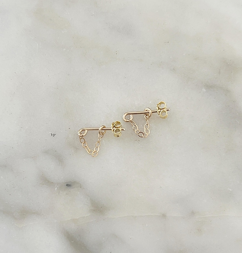 Puede incluir: Un par de pendientes de cadena dorados. Cada pendiente tiene un poste con una cadena que cuelga en forma de V. Los pendientes se muestran sobre una superficie de m&aacute;rmol blanco y gris.