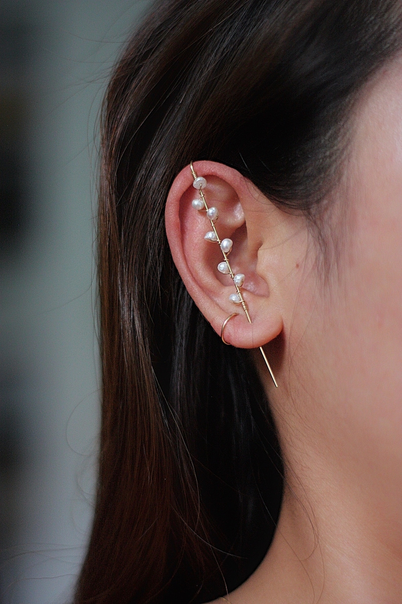 Puede incluir: Un pendiente de oreja dorado con un delicado dise&ntilde;o de peque&ntilde;as perlas blancas. El pendiente de oreja se curva a lo largo de la oreja, con una barra recta que se extiende hacia abajo. La joya se lleva en la oreja.