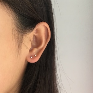 Puede incluir: Primer plano de una oreja con dos pendientes peque&ntilde;os de plata. Uno tiene forma de estrella y el otro es un simple bot&oacute;n redondo. Los pendientes est&aacute;n colocados sobre la piel de la oreja y un fondo borroso.