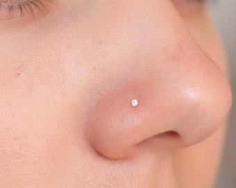 May include: Close-up of a nose with a small, sparkling, round nose stud. The stud is set in the nostril, and the skin around it is smooth and light-toned. The focus is on the jewelry.