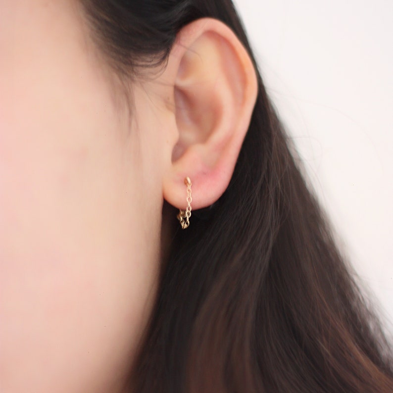 May include: A close-up of a gold chain earring worn on an ear. The earring has a small stud at the top and a delicate chain that hangs down, ending in a small cluster of gold. The background is a soft, neutral color.