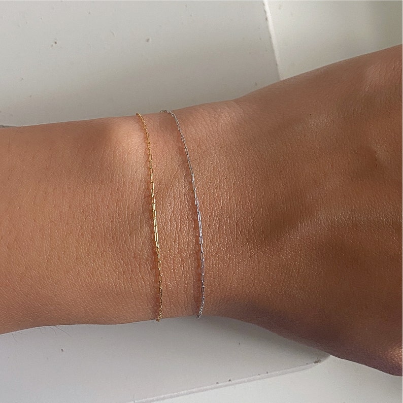 May include: Two delicate chain bracelets on a wrist. One bracelet is gold and the other is silver.
