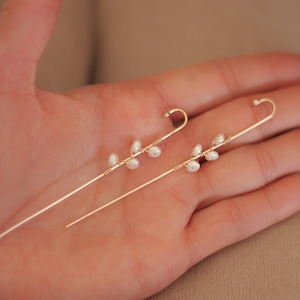 Puede incluir: Un par de trepadores de oreja dorados. Cada uno presenta un dise&ntilde;o delicado de peque&ntilde;as perlas blancas agrupadas a lo largo de un alambre fino y curvado. Los pendientes se sostienen en una mano, mostrando su estilo elegante y minimalista.