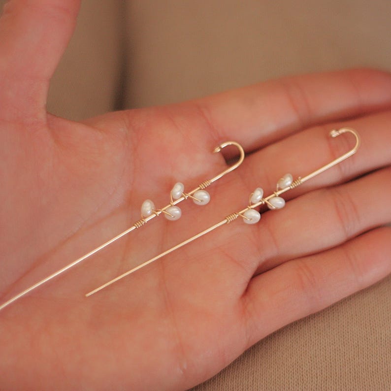 Puede incluir: Un par de trepadores de oreja dorados. Cada uno presenta un dise&ntilde;o delicado de peque&ntilde;as perlas blancas agrupadas a lo largo de un alambre fino y curvado. Los pendientes se sostienen en una mano, mostrando su estilo elegante y minimalista.