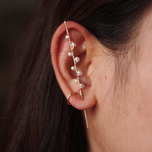 Puede incluir: Un ear cuff dorado con un dise&ntilde;o delicado con peque&ntilde;as perlas blancas. El ear cuff se curva a lo largo de la oreja, con un peque&ntilde;o pendiente de aro en la parte inferior. La joya se lleva en la oreja.