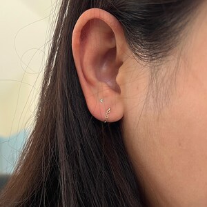 Puede incluir: Primer plano de una oreja con un peque&ntilde;o pendiente de cadena dorada y un peque&ntilde;o pendiente de bot&oacute;n. El pendiente de cadena es un bucle continuo de peque&ntilde;os eslabones conectados. El pendiente de bot&oacute;n es una peque&ntilde;a gema transparente.