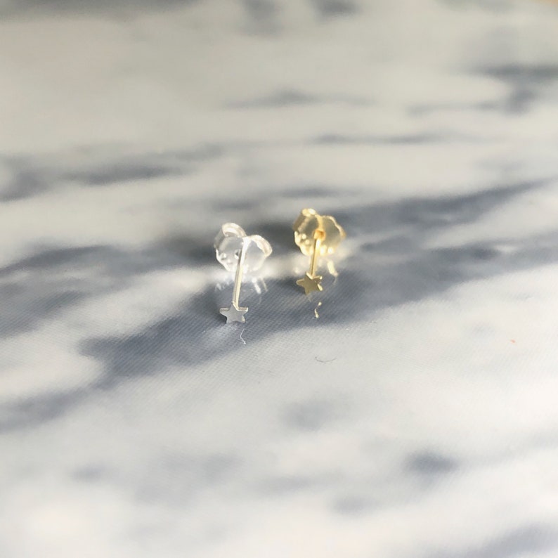 Puede incluir: Un par de pendientes de bot&oacute;n con forma de estrella. Un pendiente es de color dorado y el otro es transparente. Los pendientes se muestran sobre una superficie reflectante y veteada.