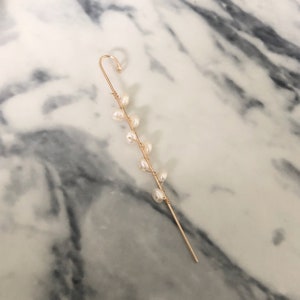 Puede incluir: Un pendiente de oreja dorado con un dise&ntilde;o delicado. Tiene un gancho curvado y un tallo delgado adornado con peque&ntilde;as perlas blancas en forma de pera. Las perlas est&aacute;n dispuestas en un patr&oacute;n lineal a lo largo del tallo.