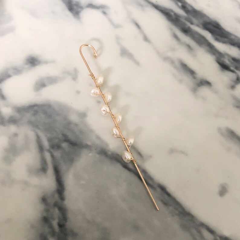 Puede incluir: Un pendiente de oreja dorado con un dise&ntilde;o delicado. Tiene un gancho curvado y un tallo delgado adornado con peque&ntilde;as perlas blancas en forma de pera. Las perlas est&aacute;n dispuestas en un patr&oacute;n lineal a lo largo del tallo.