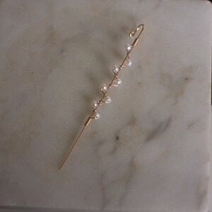 Puede incluir: Un delicado pendiente de oreja dorado con un dise&ntilde;o delgado y envuelto en alambre. Peque&ntilde;as cuentas blancas nacaradas est&aacute;n espaciadas uniformemente a lo largo del alambre, creando una est&eacute;tica sutil y elegante. El pendiente de oreja est&aacute; dise&ntilde;ado para curvarse alrededor de la oreja.