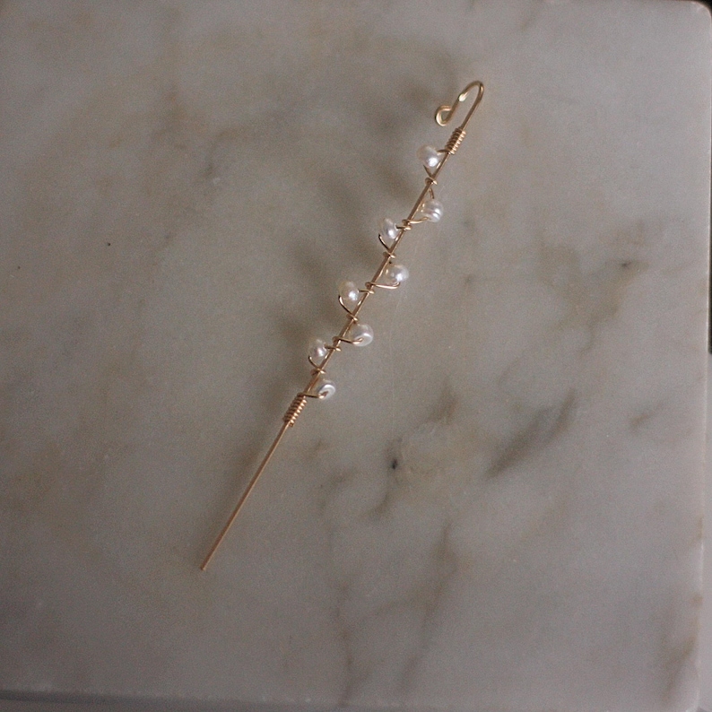 Puede incluir: Un delicado pendiente de oreja dorado con un dise&ntilde;o delgado y envuelto en alambre. Peque&ntilde;as cuentas blancas nacaradas est&aacute;n espaciadas uniformemente a lo largo del alambre, creando una est&eacute;tica sutil y elegante. El pendiente de oreja est&aacute; dise&ntilde;ado para curvarse alrededor de la oreja.