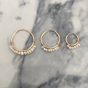 Könnte beinhalten: Drei goldene Creolen in verschiedenen Größen, jede mit einer Reihe kleiner, weißer Perlen. Die Reifen sind goldfarben, mit einer drahtumwickelten Detaillierung in der Nähe der Oberseite. Die Ohrringe werden auf einer marmorierten Oberfläche präsentiert.