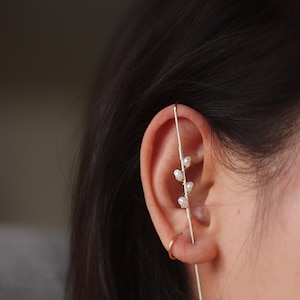 Puede incluir: Un pendiente de oreja plateado con un dise&ntilde;o delicado con peque&ntilde;as perlas blancas. El pendiente de oreja se lleva en la oreja y tiene una barra recta que atraviesa la oreja, con una barra curva que la rodea. Tambi&eacute;n se ve un peque&ntilde;o pendiente de aro dorado.