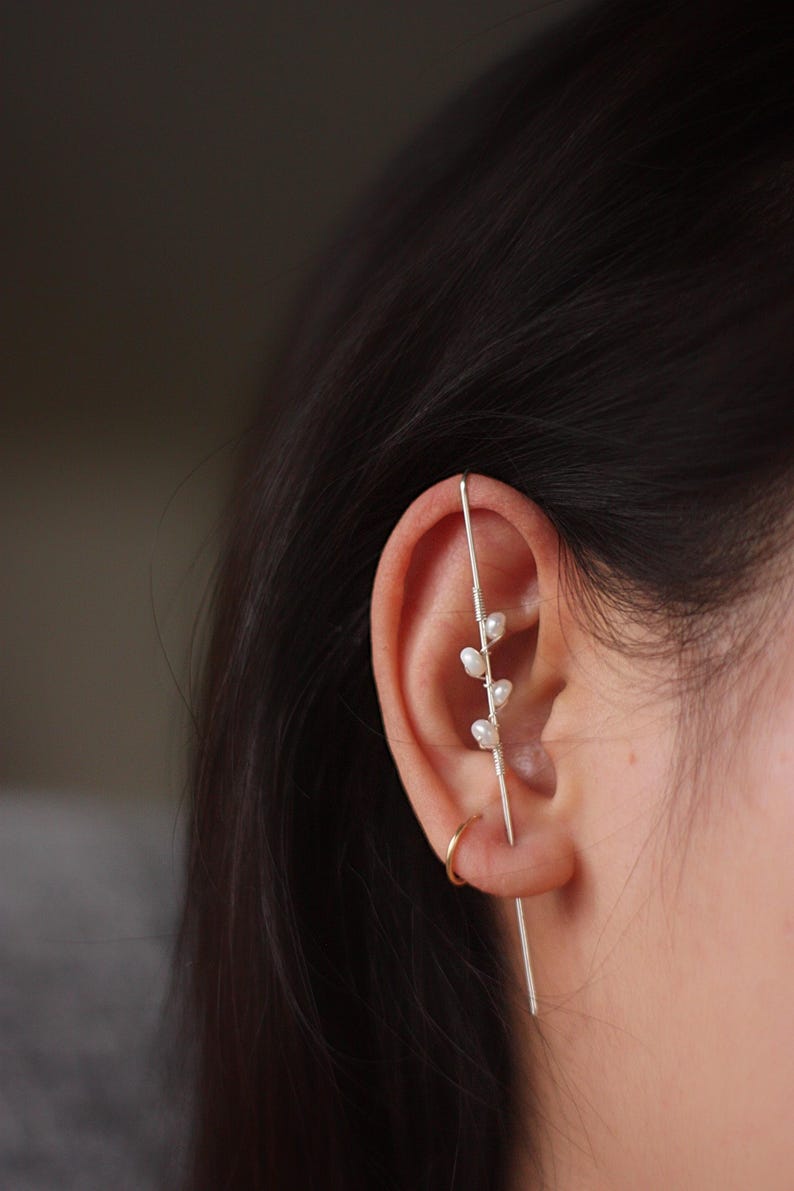 Puede incluir: Un pendiente de oreja plateado con un dise&ntilde;o delicado con peque&ntilde;as perlas blancas. El pendiente de oreja se lleva en la oreja y tiene una barra recta que atraviesa la oreja, con una barra curva que la rodea. Tambi&eacute;n se ve un peque&ntilde;o pendiente de aro dorado.