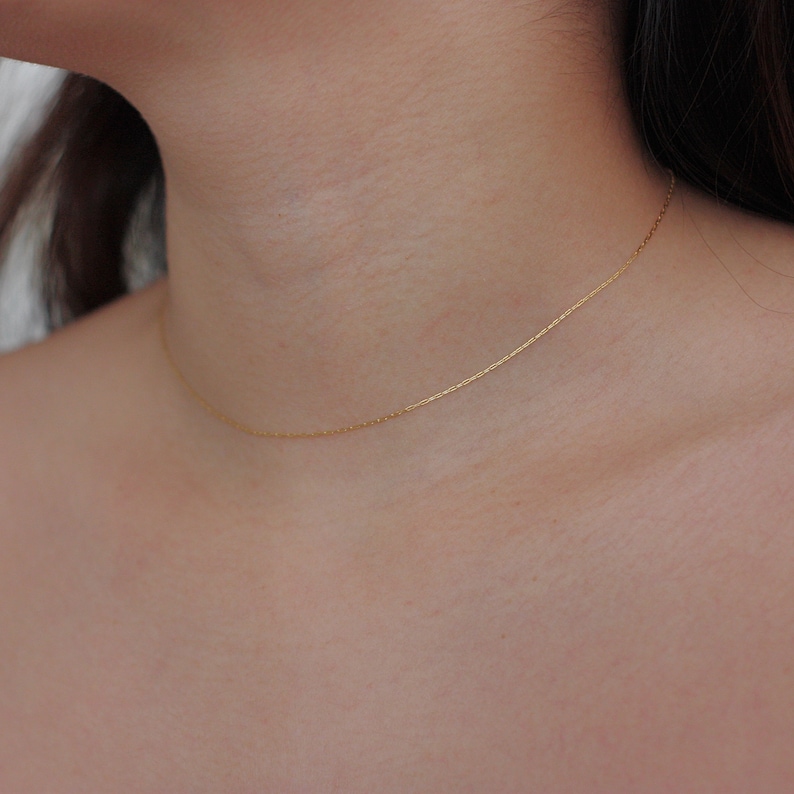 May include: A delicate gold-colored chain necklace resting on a person's neck. The necklace is thin and appears to be made of small, connected links. The background is a soft, neutral tone, highlighting the jewelry.