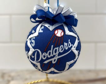 Puede incluir: Un adorno esférico de tela con fondo azul, el logotipo de los Dodgers en blanco y un gráfico de béisbol. El adorno está decorado con una superposición de tela blanca en forma de estrella y rematado con un lazo azul y blanco para colgar.