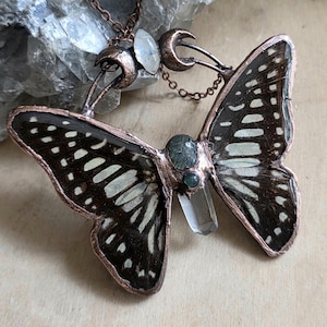 Puede incluir: Un collar con colgante de mariposa en tono cobre con una cadena. Las alas de la mariposa son negras y blancas con un patrón detallado. El colgante presenta detalles de cristal transparente y verde. El collar se exhibe sobre una superficie de madera clara.