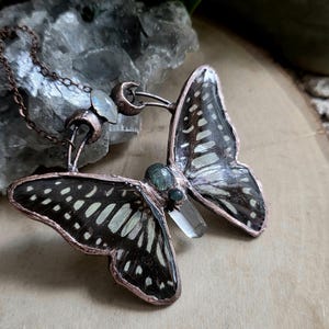 Op de afbeelding: Een vlinderhangerketting in kopertinten. De vlindervleugels zijn zwart en wit met een gedetailleerd patroon. De hanger heeft een helder kristal en kleine groene stenen. De ketting is ook koperkleurig.