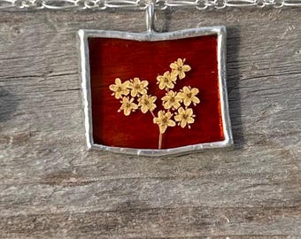 Op de afbeelding: Een zilveren ketting met een vierkante hanger met een geperst bloemenmotief. De bloem is wit en de achtergrond is rood.