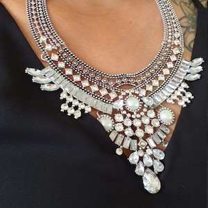 May include: Silver-toned statement necklace with clear crystals and faux pearls. The necklace features a bib design with multiple layers of sparkling stones and a chain closure. The necklace is worn over a black top.