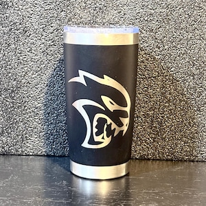 May include: Black tumbler with a silver rim and lid. The tumbler features a white graphic of a snarling cat. The tumbler is cylindrical and appears to be made of stainless steel. The background is a textured gray surface.