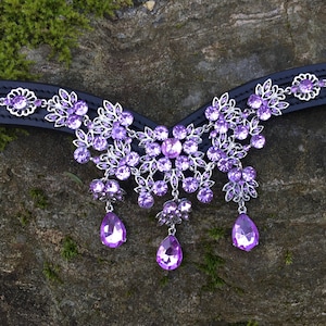 May include: A black leather horse browband with a silver and purple crystal design. The browband features a large, ornate centrepiece with dangling crystals.