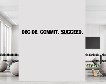 以下が含まれることがあります： 白い壁に黒い文字で「DECIDE. COMMIT. SUCCEED.」と書かれています。壁はジムの環境にあり、両側にウェイトとエクササイズ機器が見えます。