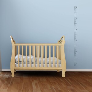Op de afbeelding: Een houten wieg met een wit matras en een witte muur met een zwarte hoogtekaart erachter. De kaart heeft nummers van 1 tot 6 voet.