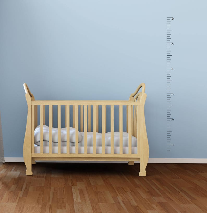 Op de afbeelding: Een houten wieg met een wit matras en een witte muur met een zwarte hoogtekaart erachter. De kaart heeft nummers van 1 tot 6 voet.