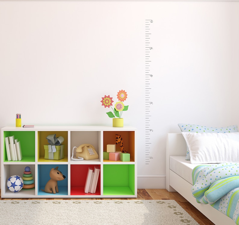 Op de afbeelding: Een witte boekenkast met kleurrijke vakken, gevuld met boeken, speelgoed en een telefoon. Een klein hondje figuur zit op een blauwe plank. Een groeimeter hangt aan de muur achter de boekenkast. De boekenkast staat in een kamer met een wit bed en een tapijt.