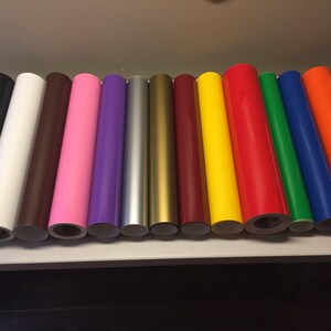 Op de afbeelding: Een verzameling van 14 rollen vinyl-kleeffolie in verschillende kleuren, waaronder zwart, wit, bruin, roze, paars, zilver, goud, rood, geel, oranje, groen, blauw en lichtblauw.