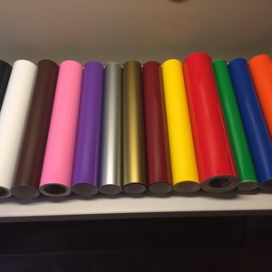 Puede incluir: Surtido de 14 rollos de adhesivo de vinilo en varios colores, incluyendo negro, blanco, marr&oacute;n, rosa, morado, plateado, dorado, amarillo, rojo, verde, azul y naranja. Los rollos est&aacute;n dispuestos en una fila sobre una mesa.