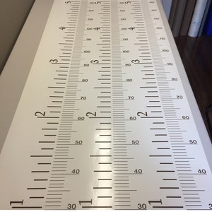Op de afbeelding: Een witte groeikaart met bruine markeringen in centimeters en inches. De kaart is gemarkeerd van 30 tot 180 centimeter en van 12 tot 72 inch.