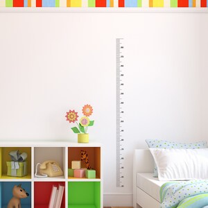 Op de afbeelding: Een witte muur met een zwarte liniaal die tot 180 cm meet, een wit rek met kleurrijke vakken, een speelgoedhond, een telefoon, een speelgoedgiraf en een bed met een blauw-wit gestreept dekbed.