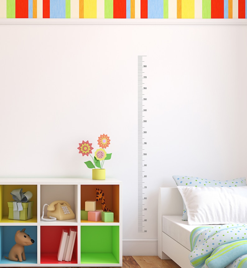 Op de afbeelding: Een witte muur met een zwarte liniaal die tot 180 cm meet, een wit rek met kleurrijke vakken, een speelgoedhond, een telefoon, een speelgoedgiraf en een bed met een blauw-wit gestreept dekbed.
