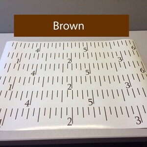 Op de afbeelding: Een witte liniaal met zwarte markeringen, die inches aangeeft. De liniaal is gelabeld met de nummers 1, 2, 3, 4, 5 en 6. Het woord "Brown" staat in bruine tekst op een bruine rechthoek boven de liniaal.