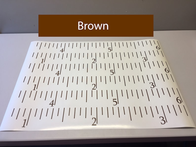 Op de afbeelding: Een witte liniaal met zwarte markeringen, die inches aangeeft. De liniaal is gelabeld met de nummers 1, 2, 3, 4, 5 en 6. Het woord "Brown" staat in bruine tekst op een bruine rechthoek boven de liniaal.