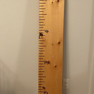 Op de afbeelding: Een houten groeimeter met zwarte markeringen en nummers van 1 tot 6. Op de meter staan de woorden "Daddy" en "Mommy" geschreven.