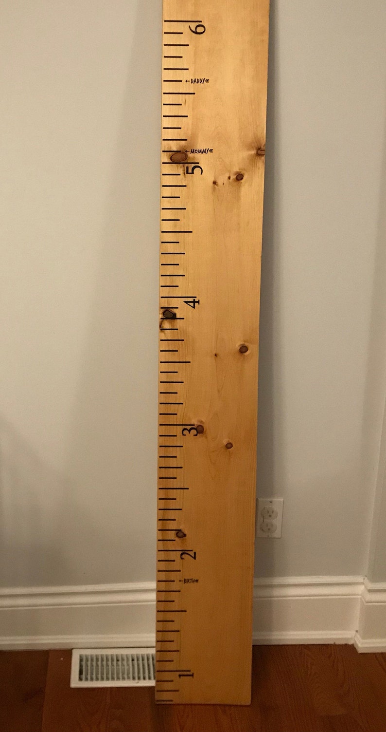 Op de afbeelding: Een houten groeimeter met zwarte markeringen en nummers van 1 tot 6. Op de meter staan de woorden "Daddy" en "Mommy" geschreven.